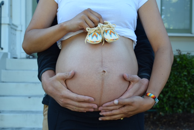 Angel holds a tiny pair of yellow moccasins atop her exposed pregnant belly, as she and her husband hold Angel's belly.