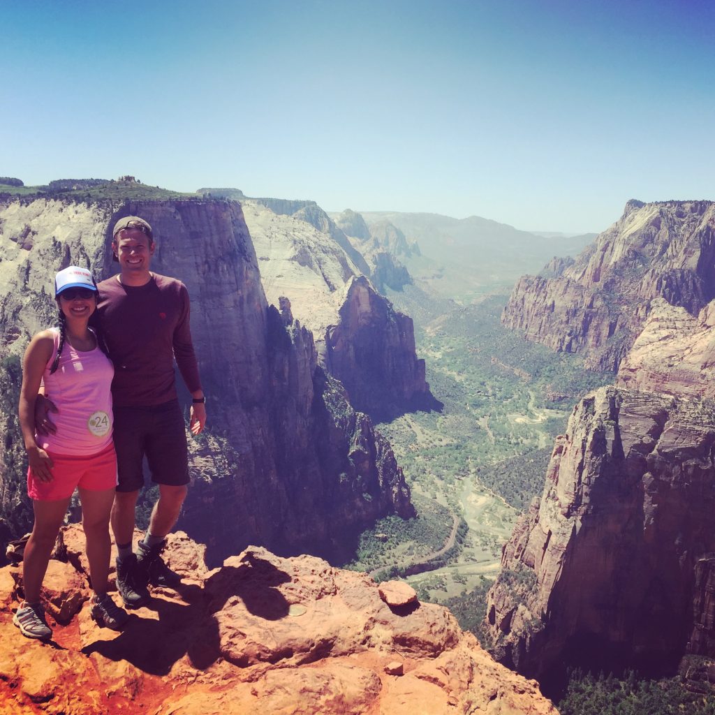 Angel and her partner smile while standing atop Angel's Landing at Zion National Park, with a view of the canyon behind them.