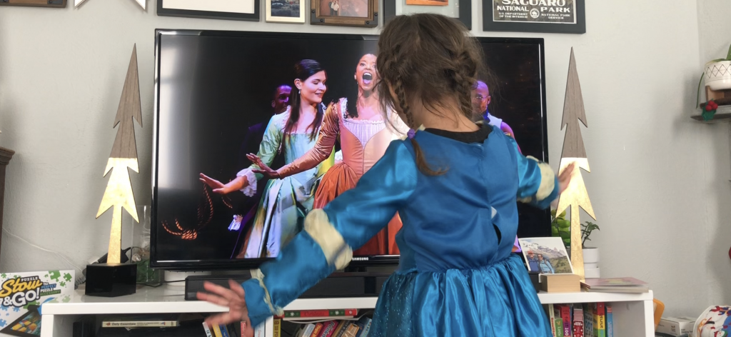 child in a formal dress dancing in front of a television showing a scene from Hamilton
