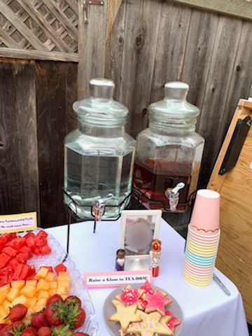 Two drink containers full of water and tea on a table next to a stack of pink, blue, and yellow paper cups, star cookies, and fruit.
