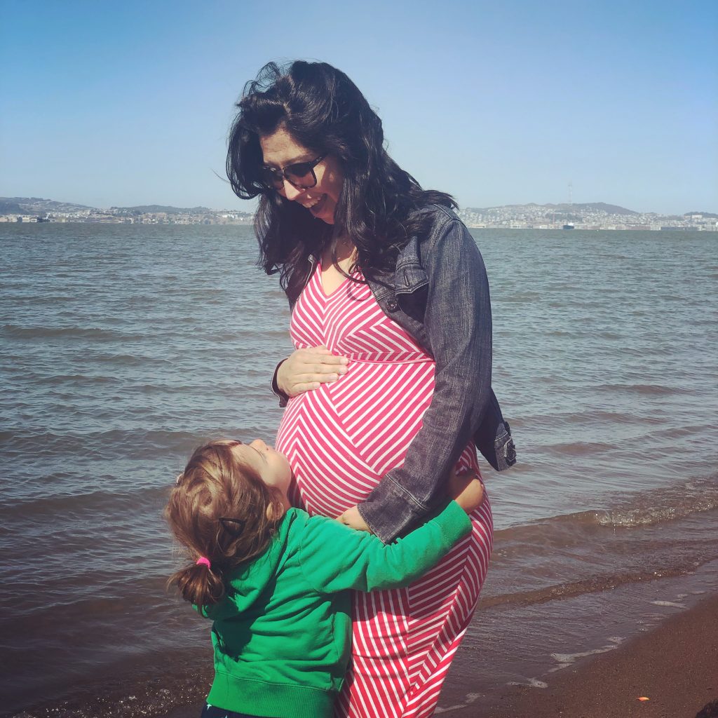 Angel looks excited in a red dress as her child hugs Angel's pregnant belly at the beach.