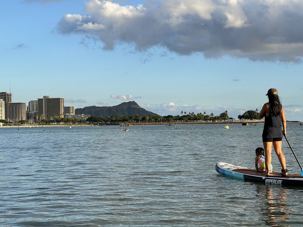 Jelly looks out towards Diamond Head while paddleboarding with her child at Ala Moana Beach Park in Honolulu, Hawaii.