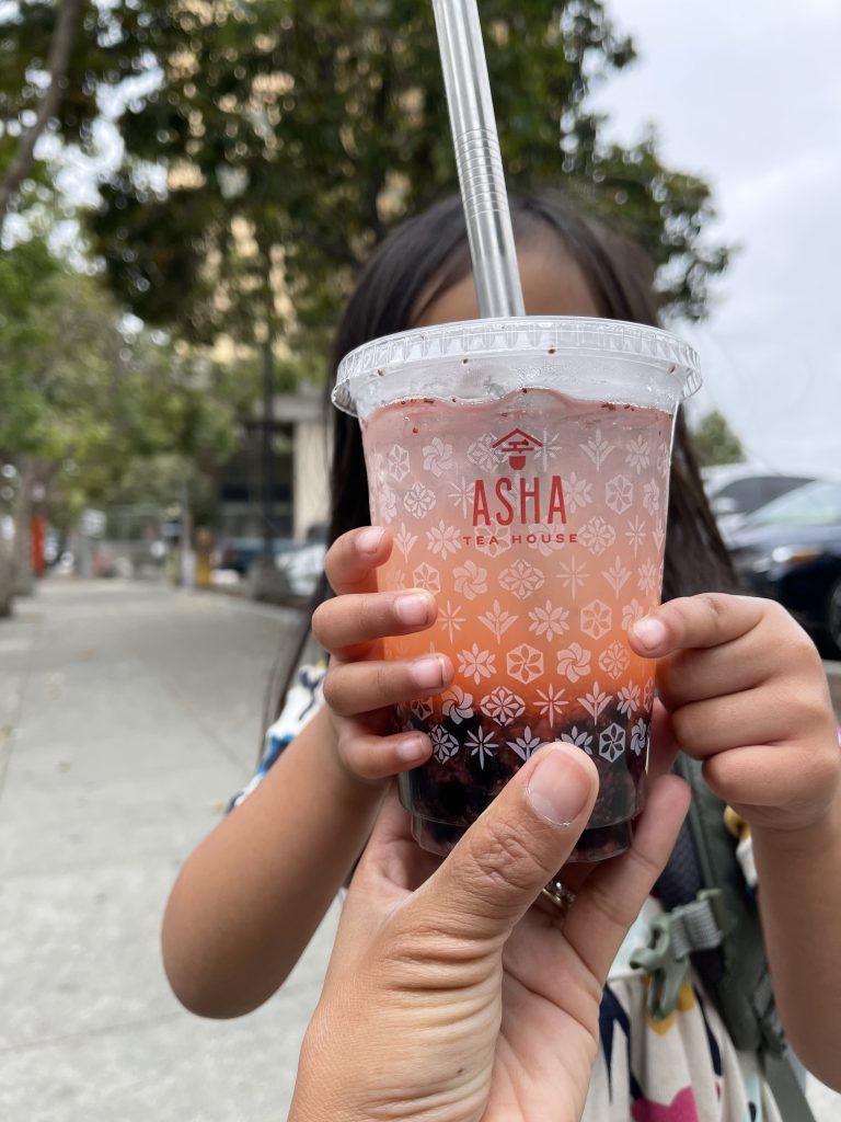 A child holds up a pink boba drink from Asha Tea House in front of their face.