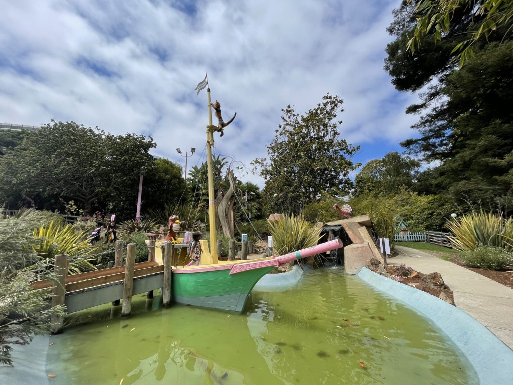 A green ship in a green lake is set against a blue-yet-cloudy sky. A child is walking atop the ship.