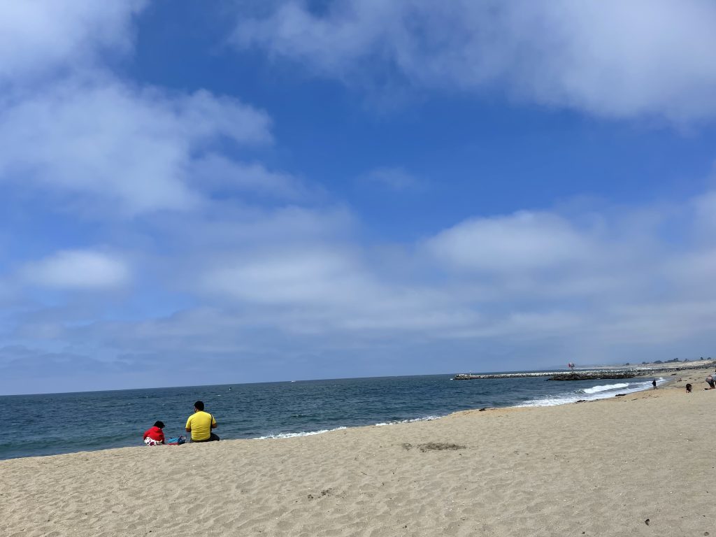 parent and child sitting on big sandy beach under blue sky with clouds