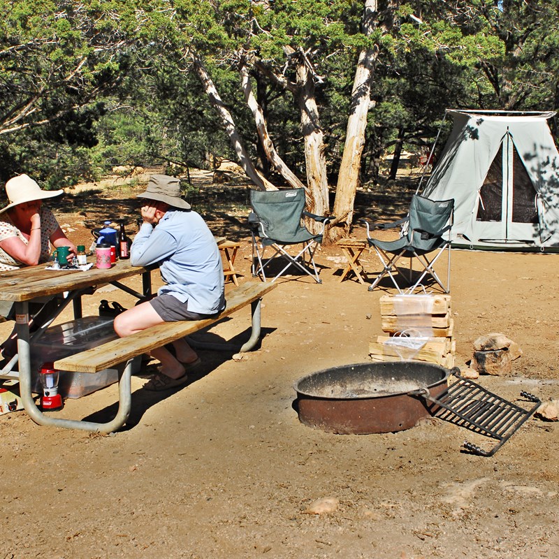 Two campers sit at a picnic table near a fire ring, camping chairs, and their tent.