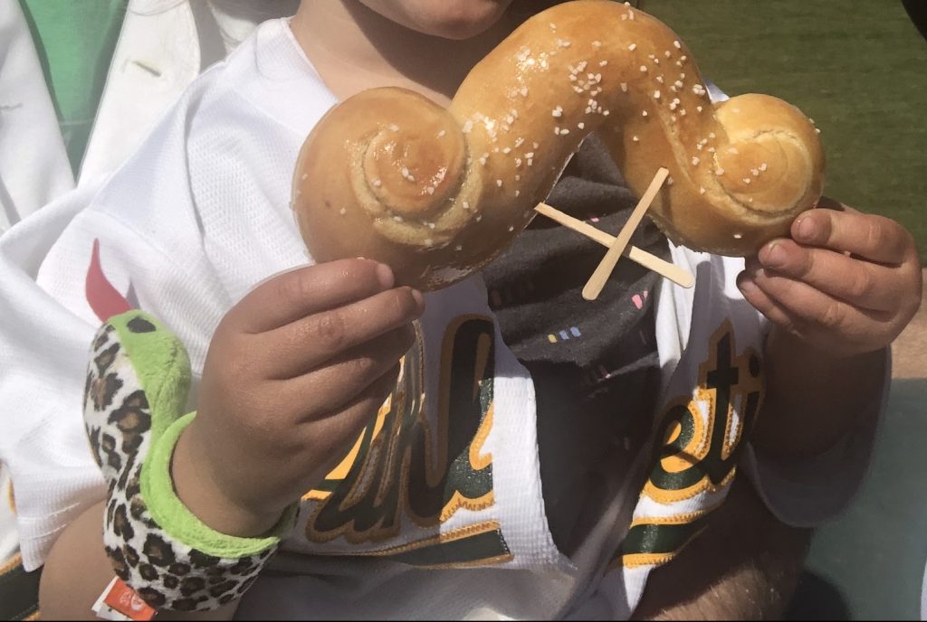 A child holding a pretzel shaped like a mustache at Hohokam Stadium for Spring Training