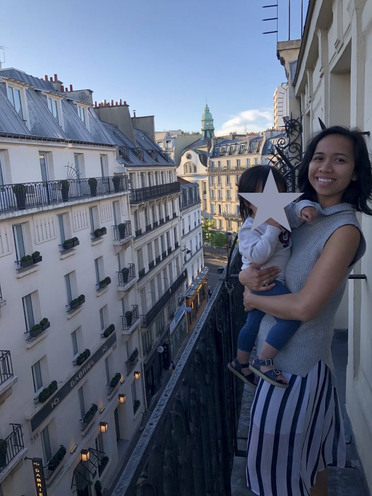 Jelly holds her child on their hotel balcony, with a view of Parisian buildings in the background.
