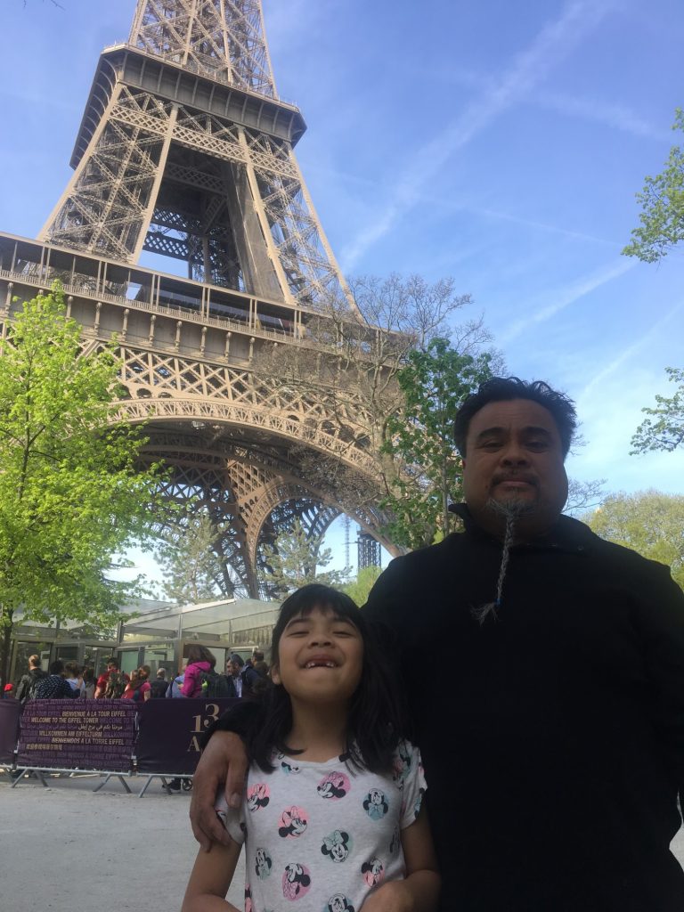 A young girl and her father smile at the camera as they stand at the base of the Eiffel Tower, which stands in the background.