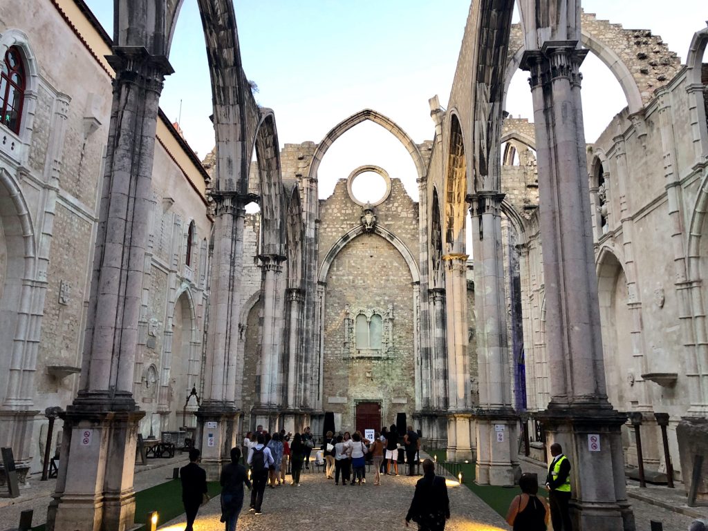 The stone columns of a gothic building with no roof impose above a crowd of visitors