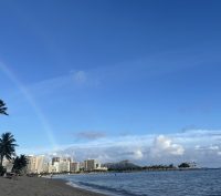 The Honolulu skyline rises above Ala Moana beach where a rainbow streaks across the sky.