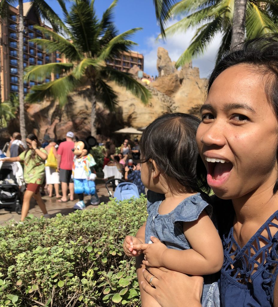 Angelica looks excitedly at the camera while holding her toddler--Mickey Mouse in an aloha shirt in the background.