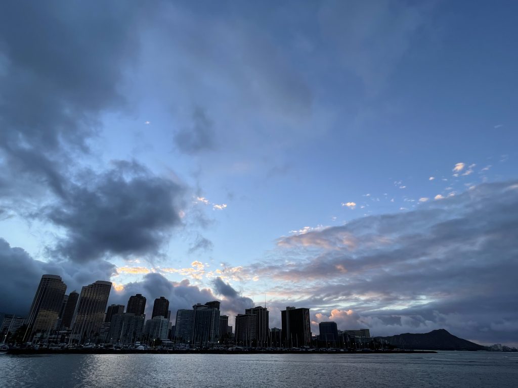 The silhouette of the Waikiki skyline stands above the water against a dark blue sky full of clouds.