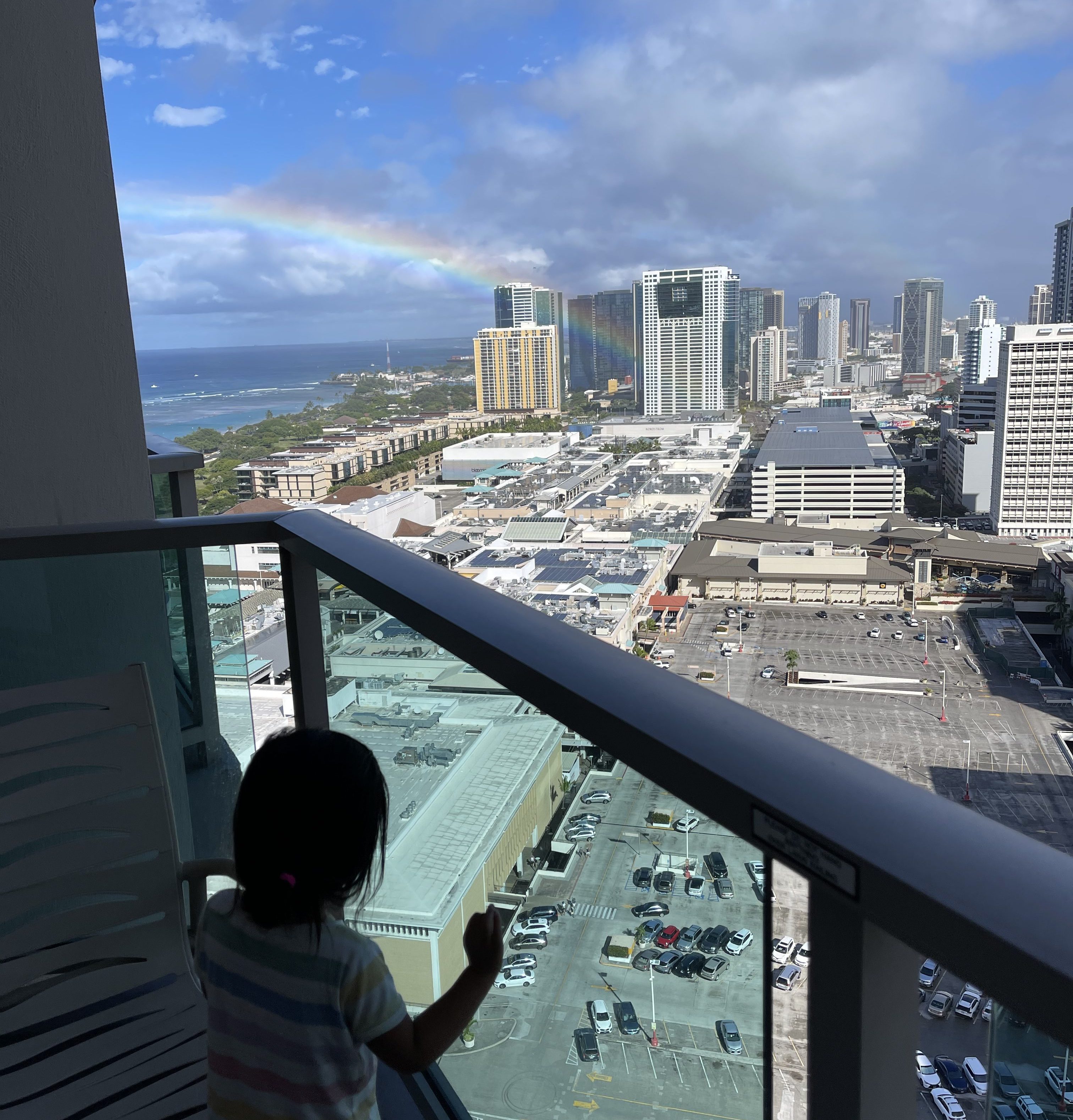 A child looks out of a glass-walled balcony to look at a rainbow that hangs above the buildings of Ala Moana.