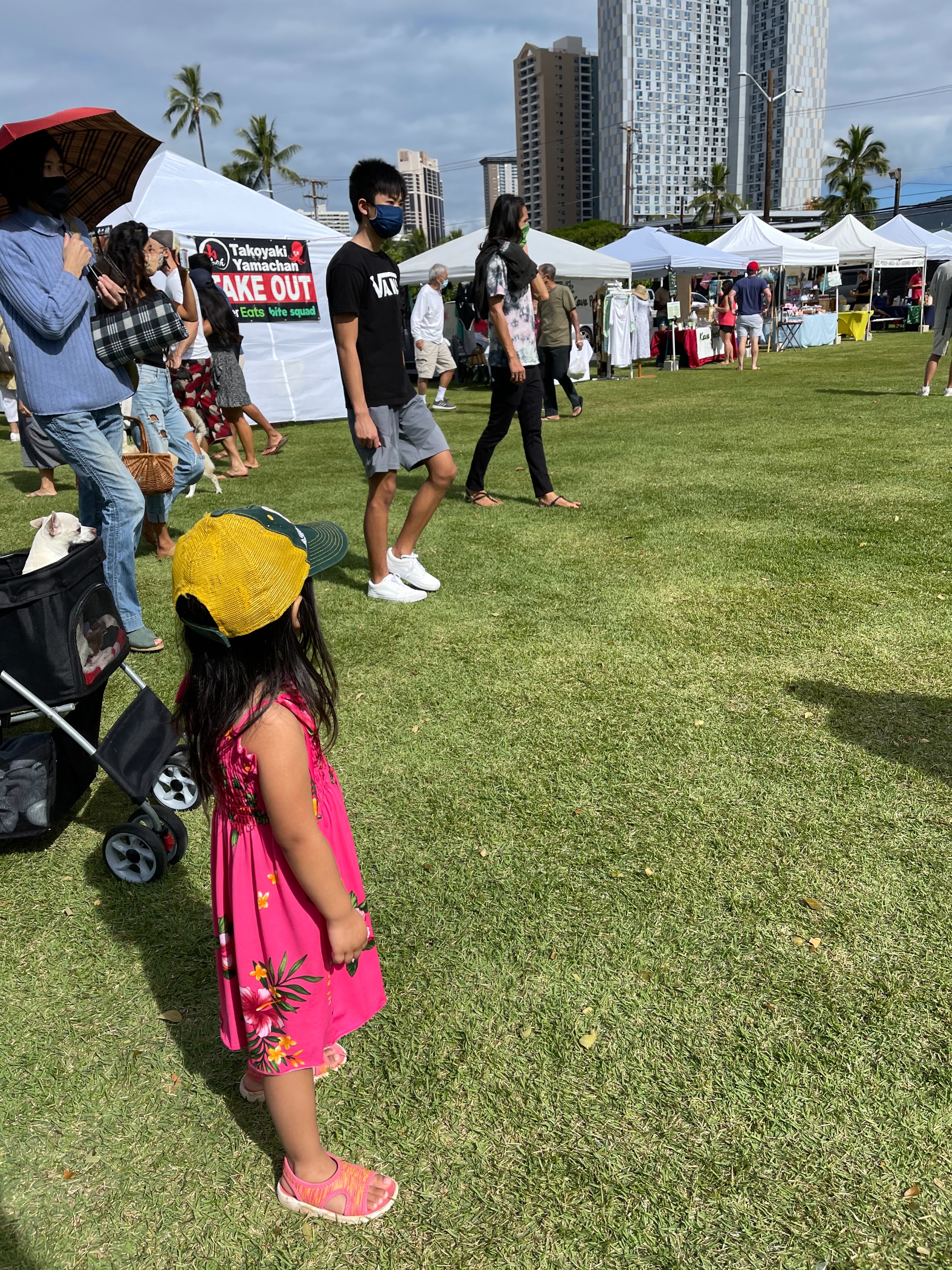 A child in a pink dress and yellow hat looks out into a field of farmers market tents and booths.