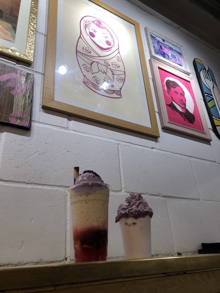 Two multi-colored and purple drinks sit on a shelf under Pilipinx-inspired artwork.