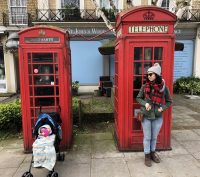 Angelica and her child (sitting in a stroller) each stand in front of two red telephone booths in London.