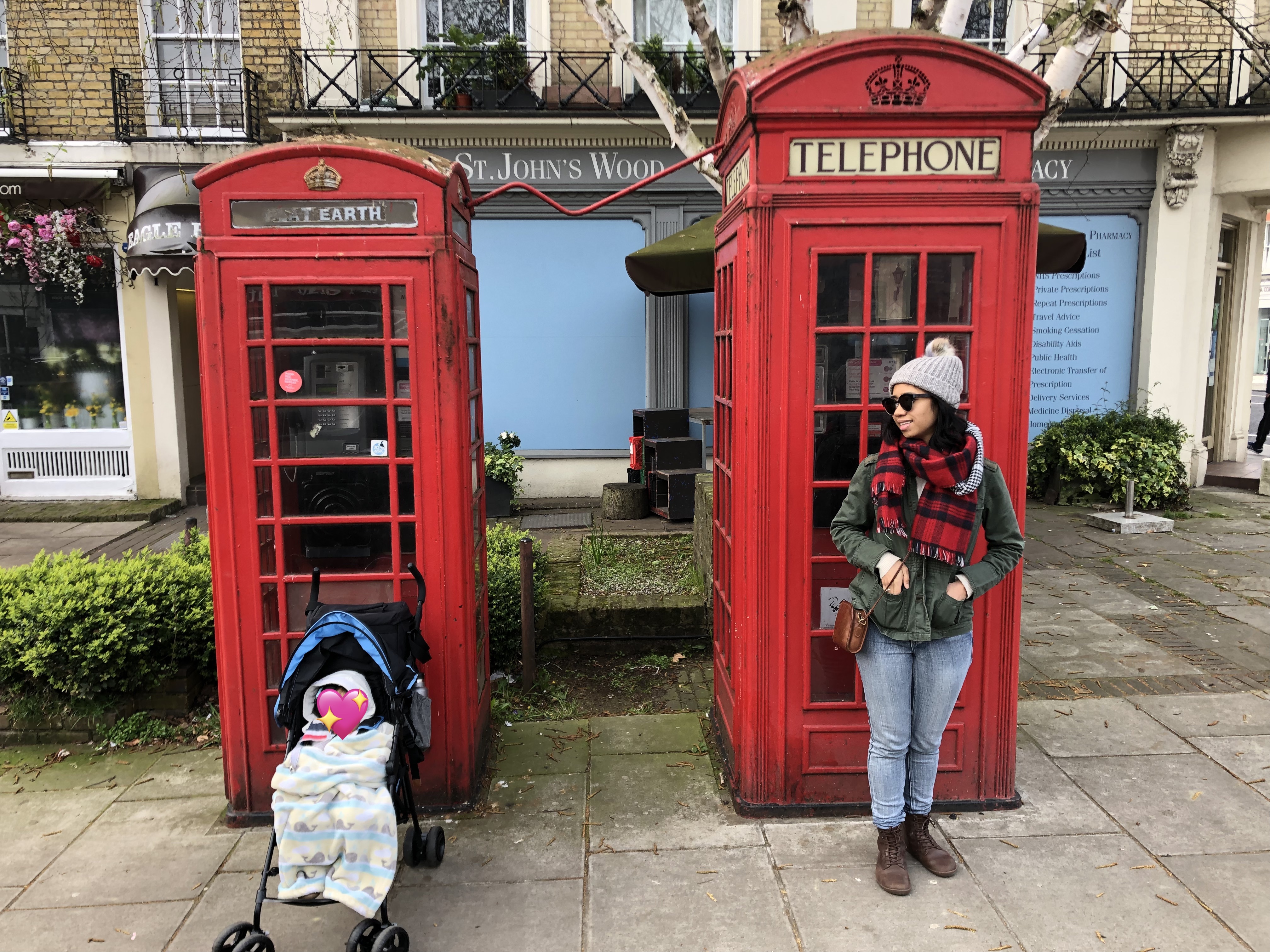 Angelica and her child (sitting in a stroller) each stand in front of two red telephone booths in London.