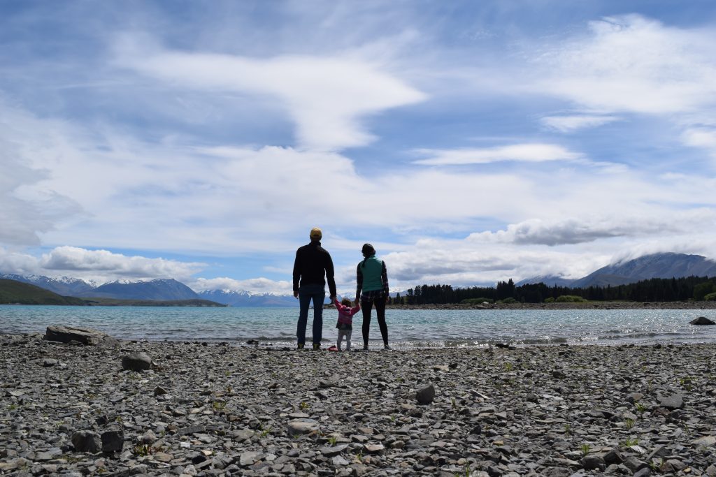 A family with a small child standing on a rocky shoreline of Lake Tekapo, New Zealand