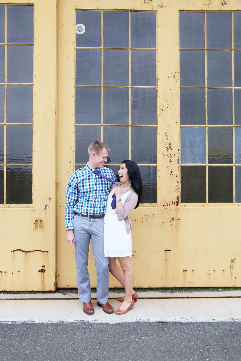 Two adults looking at each other, smiling in front of a rusty yellow building with large windows at Alameda Point in California