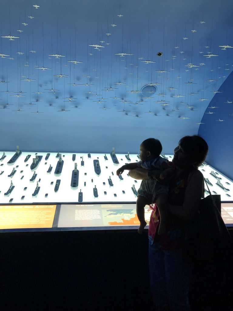 The silhouette of a baby and a woman are in the foreground of a bright display of mini naval ships.