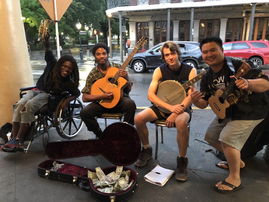 Four men sit in a row on a sidewalk holding instruments and posing for the camera