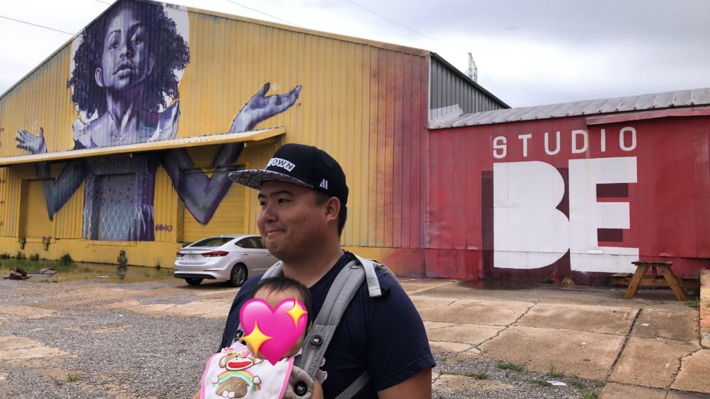 Angelica's partner holds their baby in a carrier outside of Studio Be, where a mural of an African American girl holding open her arms is painted.