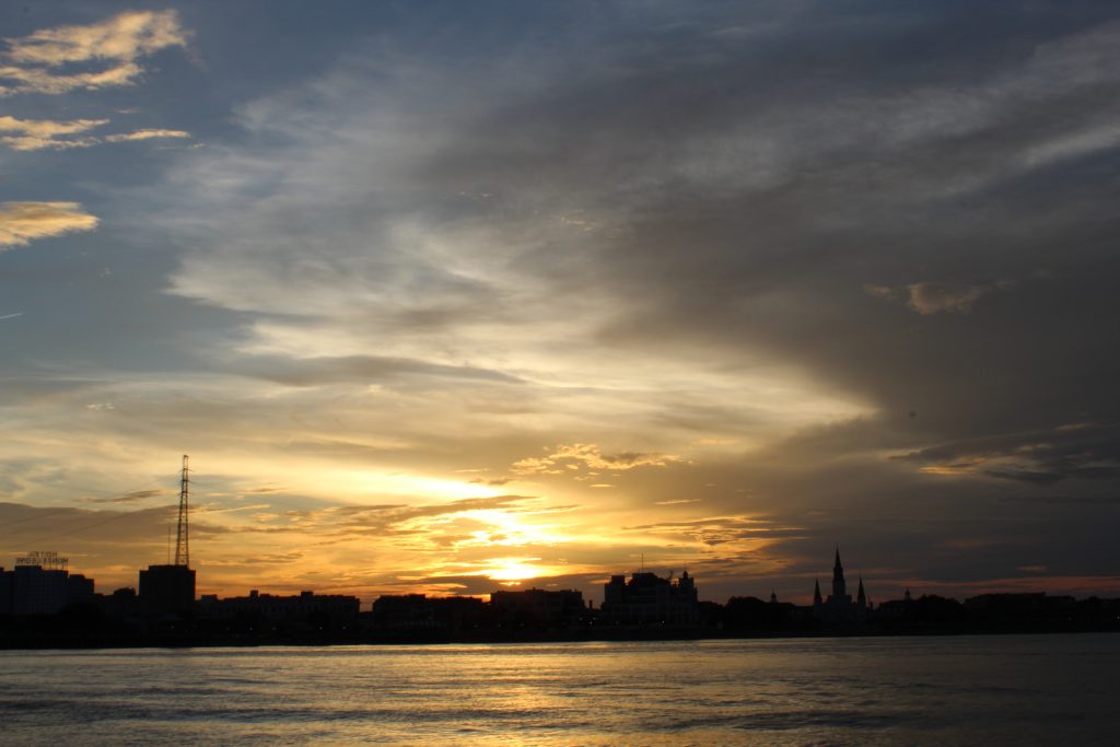 The sunsets behind the buildings of New Orleans with the river in the foreground and a darkening cloudy sky in the background.