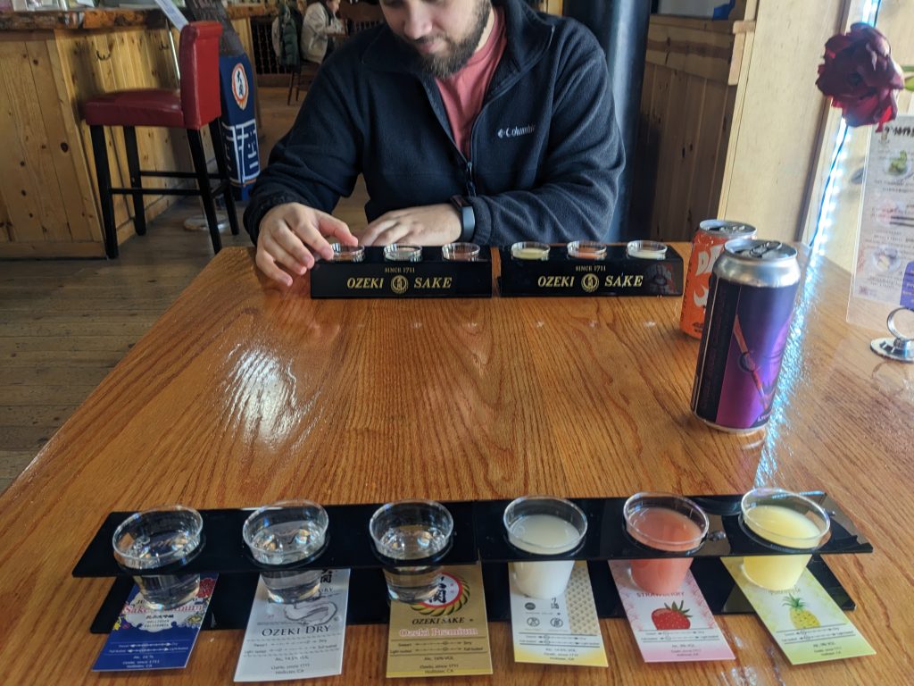 Six colorful shots of sake laid out on a table, across from which Krista partner sits.