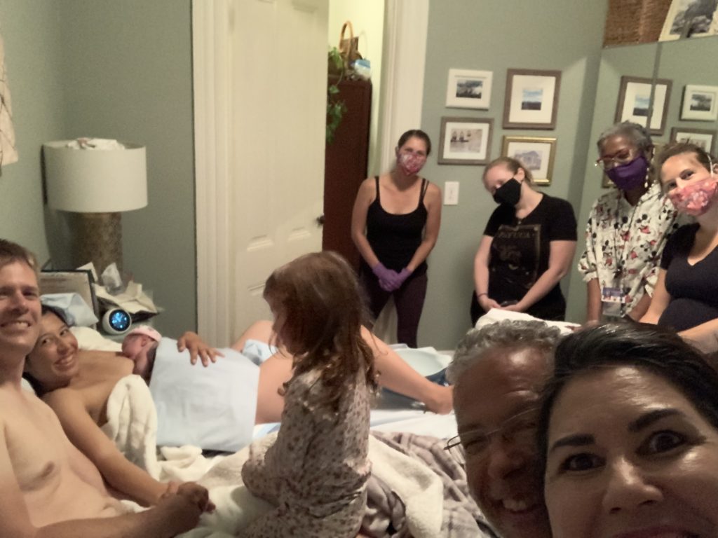 The home birth support team surrounding a woman post-delivery with a newborn in her bed