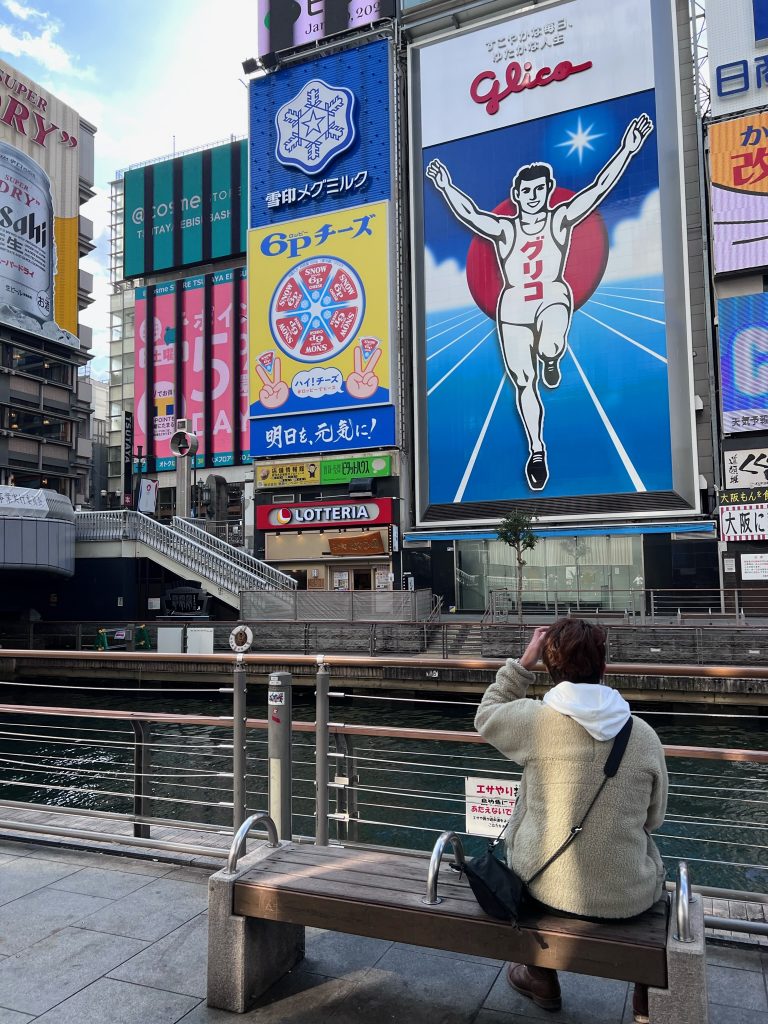 A person sits along the Dotonbori River looking out at the bright blue Glico and other ads across the river.