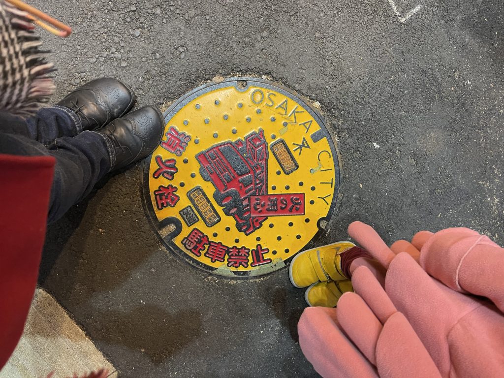 A bright yellow manhole cover is adorned with red kanji and flanked by two sets of shoes.