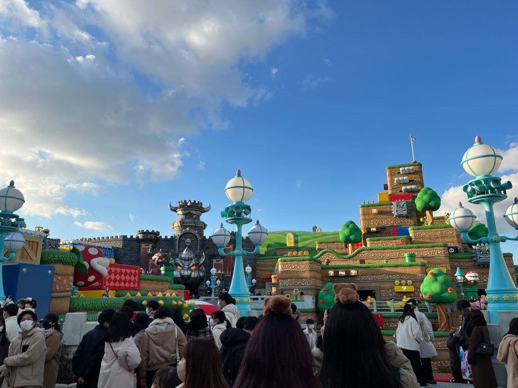 Against a bright blue and slightly cloudy sky, the structures of Super Nintendo World are seen behind a crowd of people.