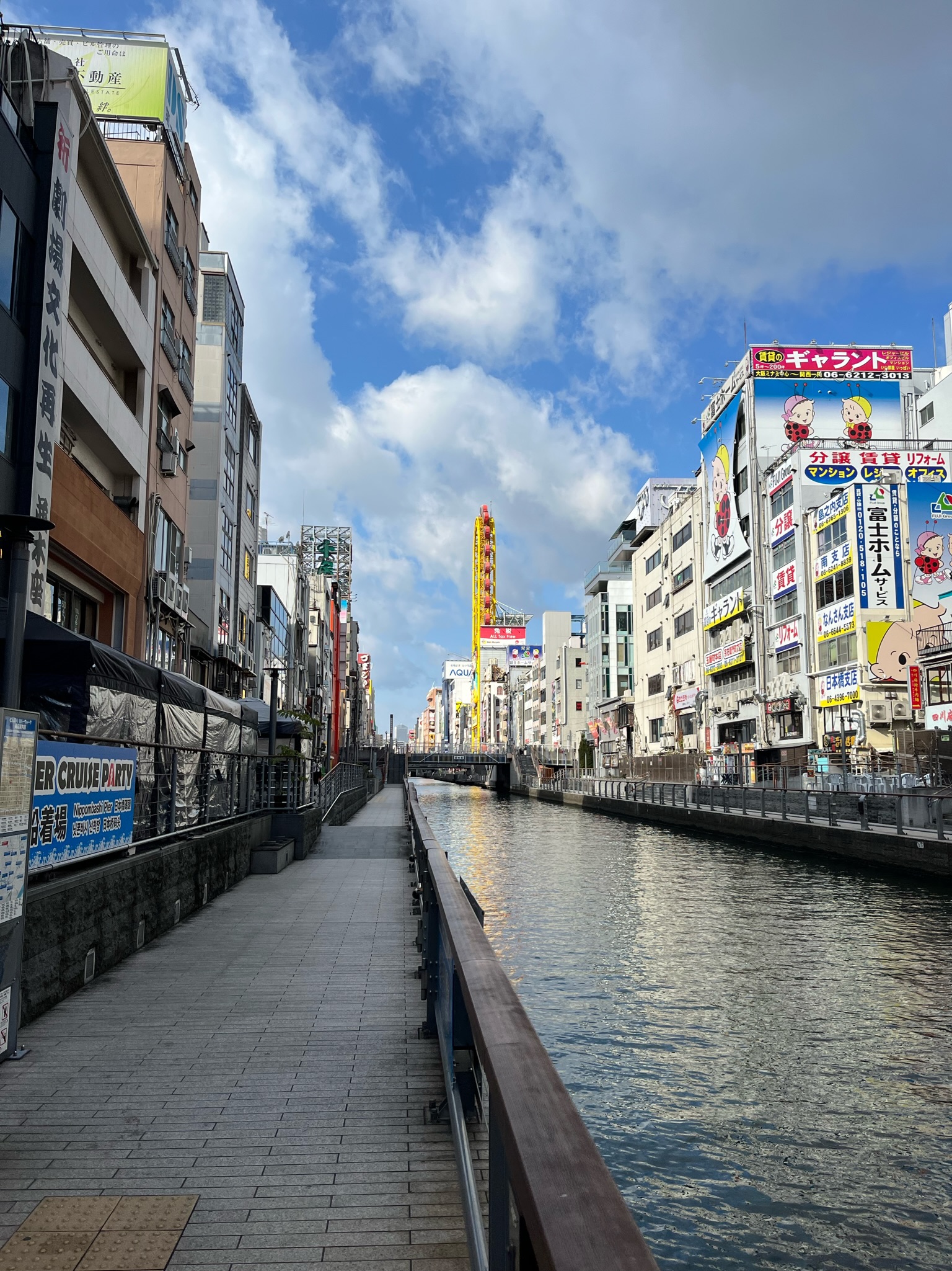 A road and the Dotonbori River are seen adjacent to one another and flanked by buildings on a slightly cloudy day.