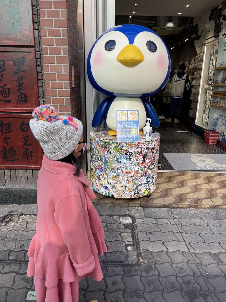 A child in a pink jacket and grey beanie looks at a cartoonish blue penguin statue in front of a storefront.