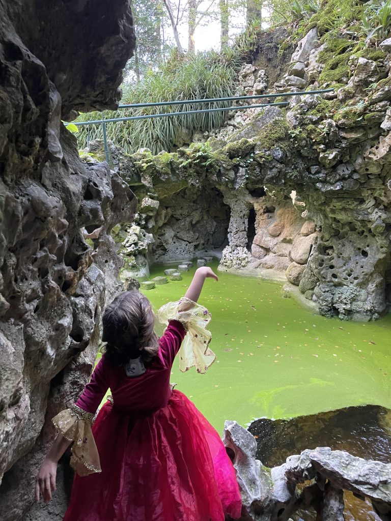 Child in a princess dress pointing to a grotto with green water at the Quinta da Regaleira in Sintra, Portugal