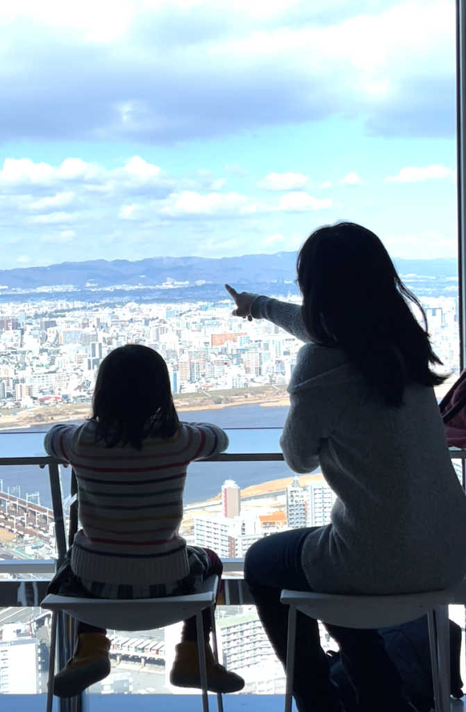 A child and her mother are silhoutted against a glass backdrop of a daytime city view.