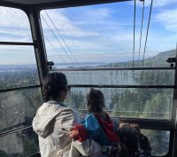 An adult and two children looking at the view of blue skies and green mountainside from the gondola up to Grouse Mountain in Vancouver BC