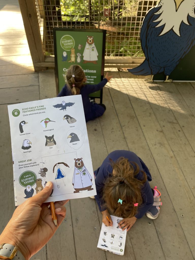 A hand holding a scavenger hunt list on paper above a child doing the hunt at Capilano Suspension Bridge Park in Vancouver