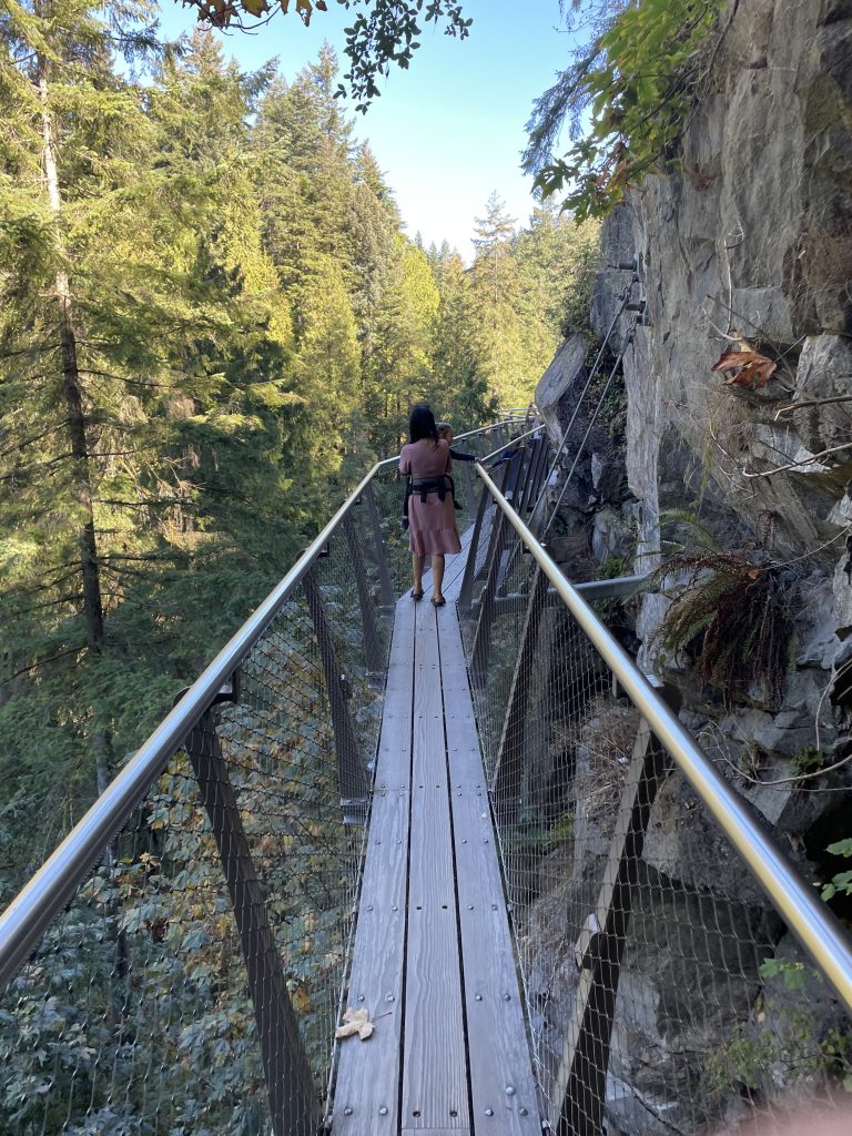 A women wearing a tushbaby carrier walking across the cliff walk of Capilano Suspension Bridge Park in Vancouver