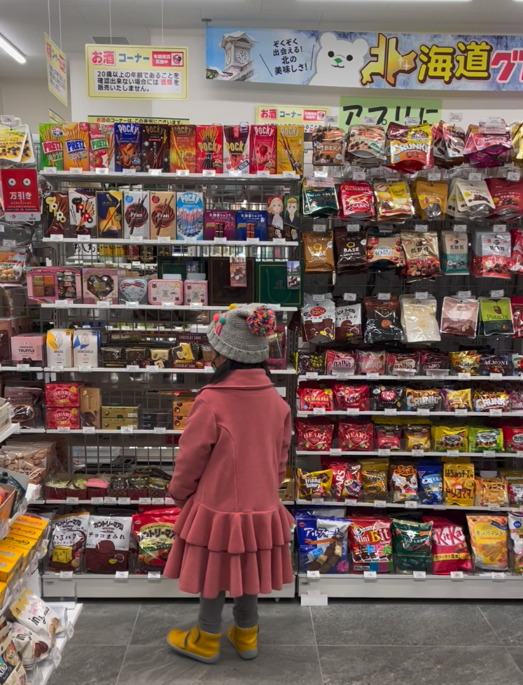 A child wearing a pink jacket, yellow shoes, and a grey beanie looks up at a colorful shelf twice her height that is full of snacks and candies at 7-11 in Tokyo.