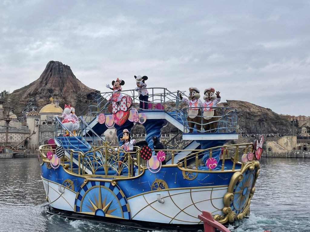 The classic Disney characters—Minnie, Mickey, Donald, Daisy, Goofy, and Chip & Dale, wave from a blue and white boat, with a volcano looming in the background.