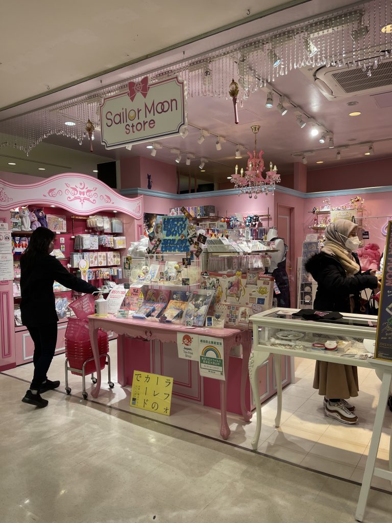 The storefront of the Sailor Moon Store dons a large sign hanging from the ceiling, and lots of knick knacks on pink shelves.