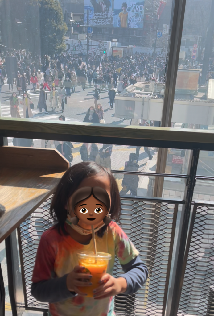 A child wearing a tie-dye shirt and holding an orange juice stands in the foreground, in a stairwell that overlooks a crowd of pedestrians at Shibuya Crossing on a sunny day.