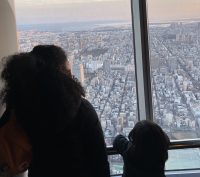 A woman and a child are silhouetted as they look out large windows that overlook the city of Tokyo during sunset.