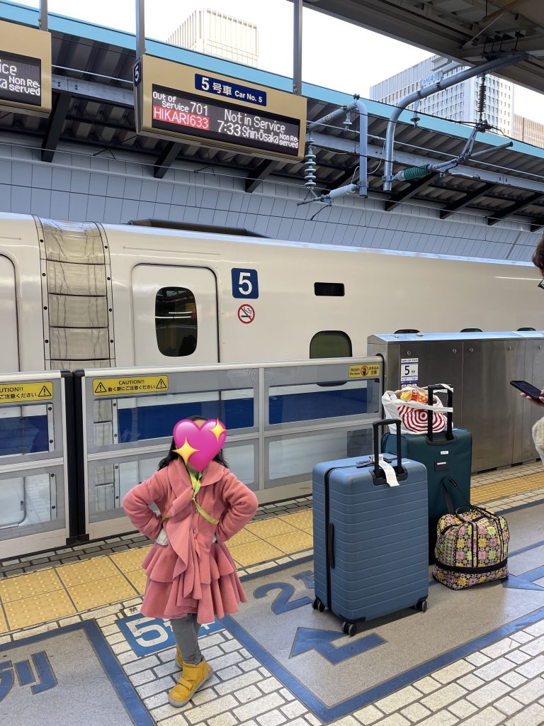 A child wearing a pink jacket makes a heart with her hands as she stands next to a pile of luggages in front of a train car in Tokyo.