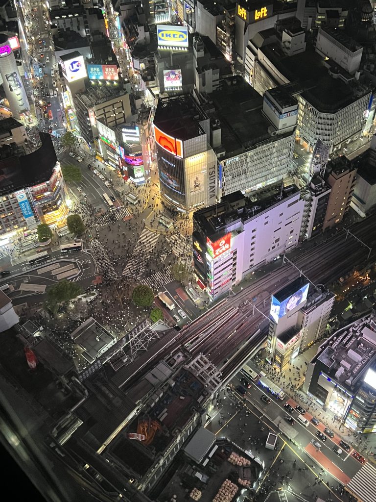 A bird's eye view of brightly lit city highrises and large streets and crosswalks filled with pedestrians.