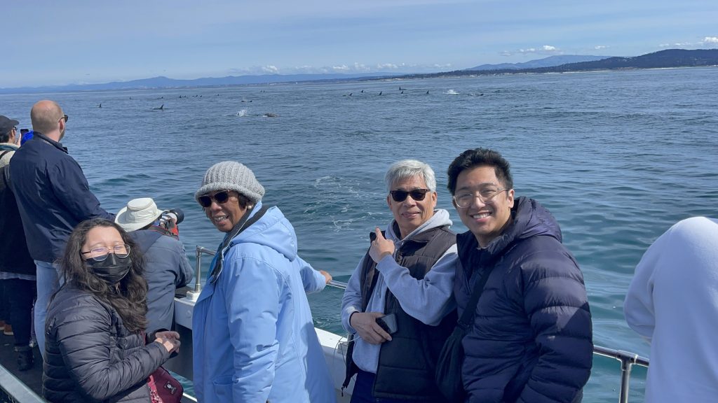 Four adults smile at the camera as they stand in the back of a boat with the Monterey Bay and breaching Risso's dolphins in the background!