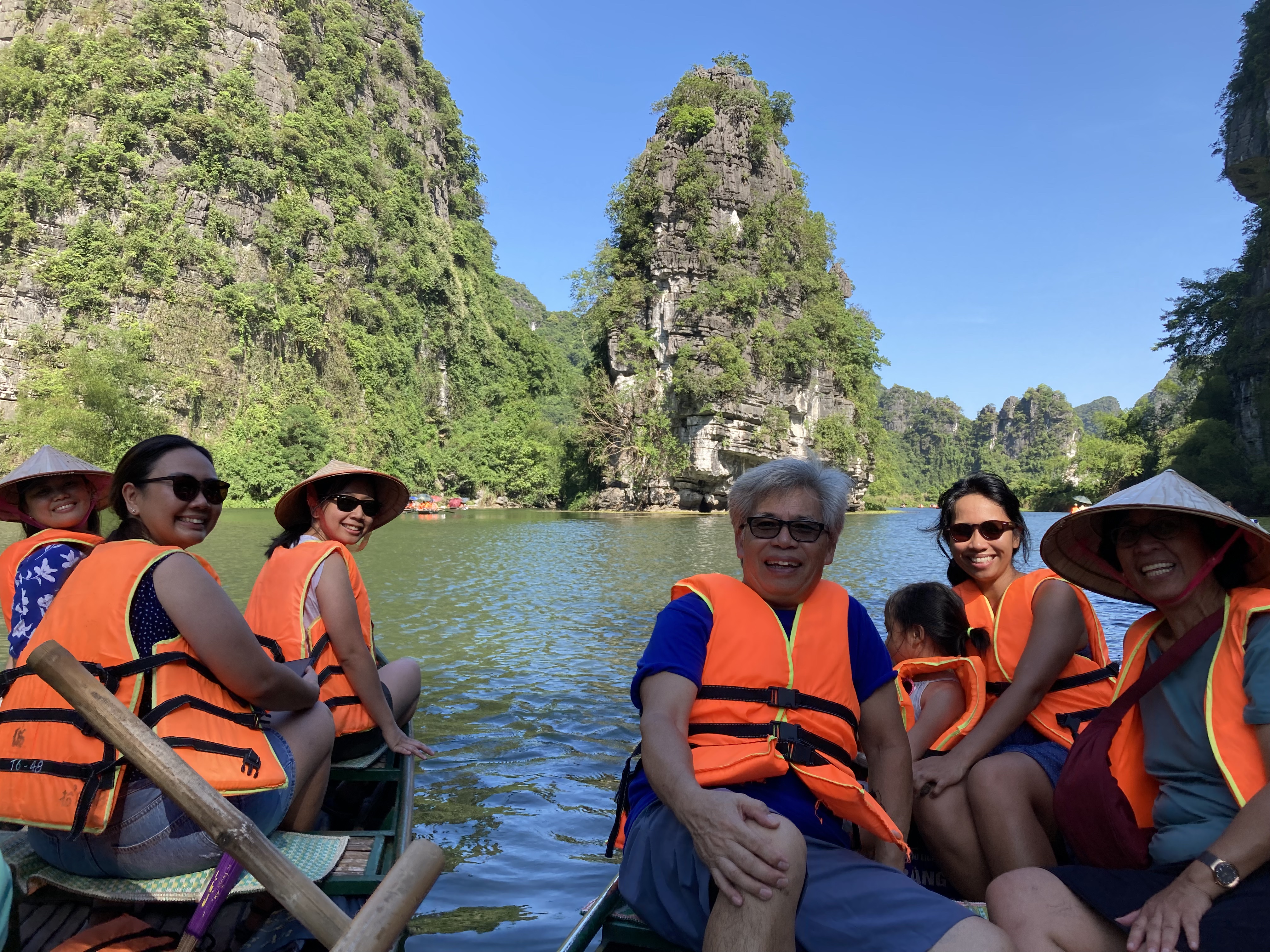 Family members donning orange life vests smile at the camera from their seats in two rowboats, with green karsts in the background.