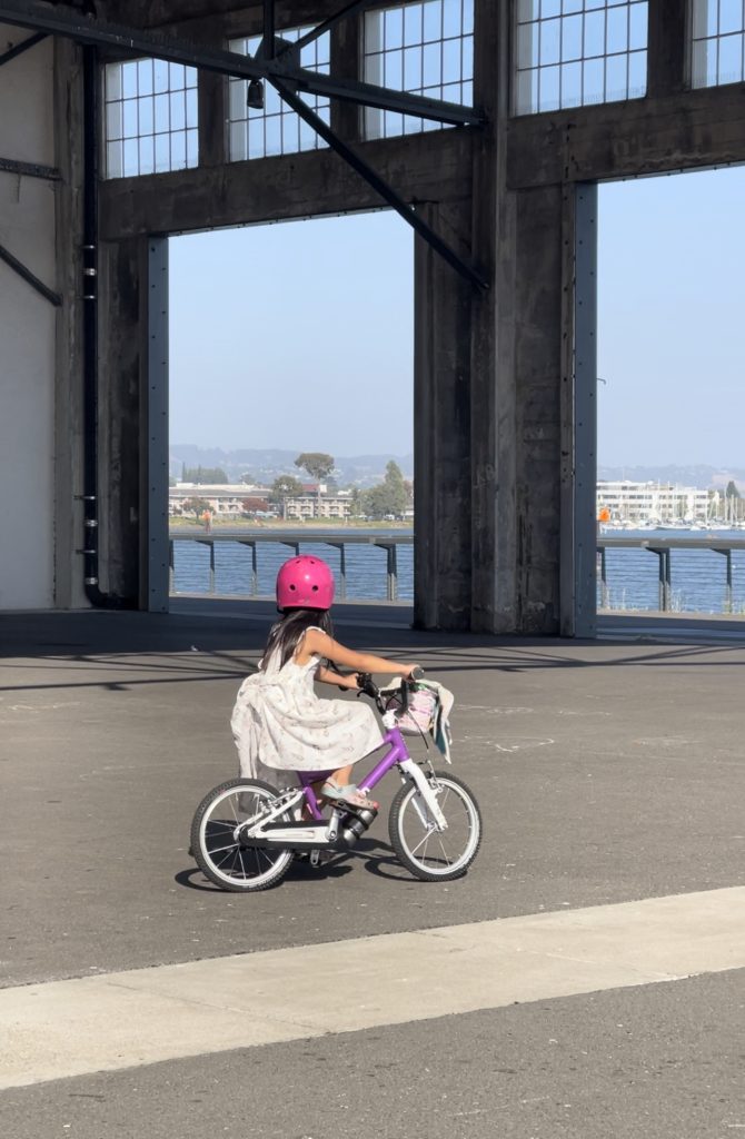 A child in a light pink dress and hot pink helmet rides a purple bike on concrete with the Oakland estuary in the background.